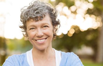 Smiling person with short, curly gray hair stands outdoors in soft lighting, wearing a light blue shirt.